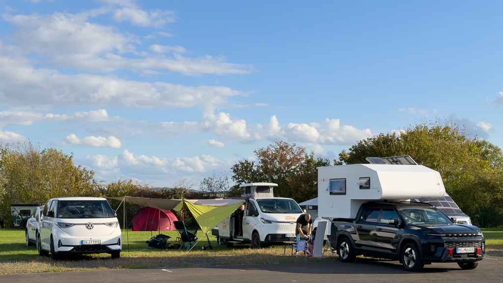 A camping scene featuring three vehicles: a white electric car, a black pickup truck, and a white camper van. There are tents and camping gear set up in a grassy area under a blue sky with fluffy clouds.