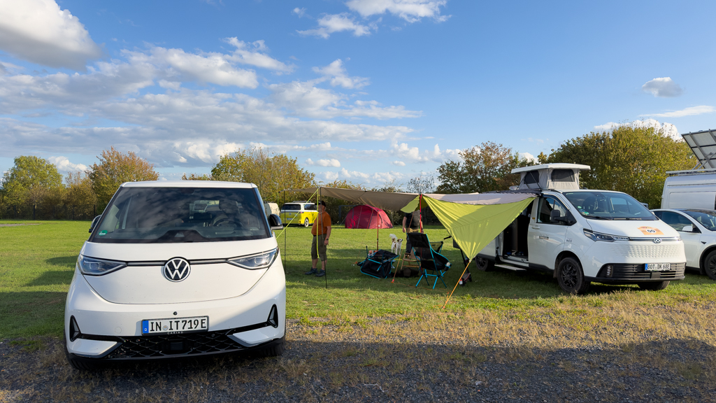 A white Volkswagen electric vehicle parked in a campsite, alongside a white camper van and a green tarp shelter. Two people are seen near chairs, with tents and trees in the background under a partly cloudy sky.