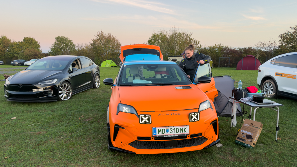 A bright orange Alpine electric car is parked with its hood open, while a person leans in to check something inside. In the background, a black Tesla Model X and a white SUV are visible, with camping tents set up on a grassy field under a clear sky.