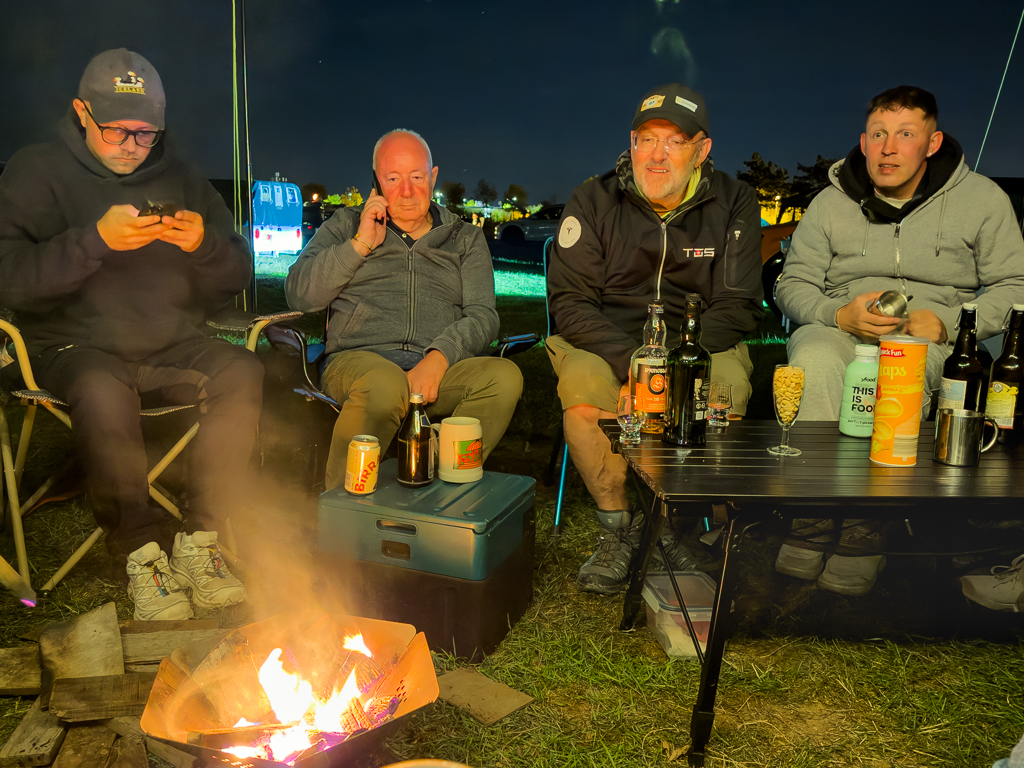 Group of four men sitting around a campfire at night, enjoying drinks and snacks. One man is using a smartphone, while others are engaged in conversation and relaxing.