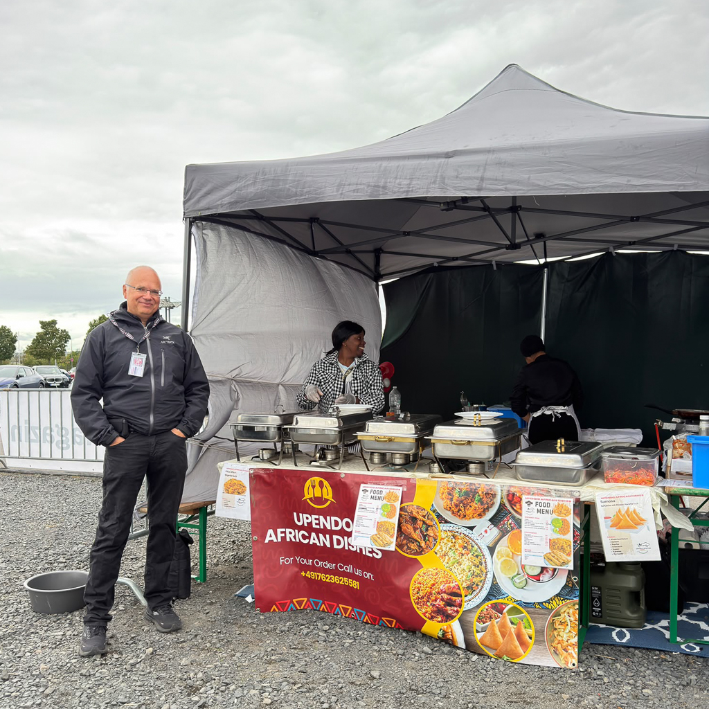 A man smiling in front of a food stall called 'Upendo African Dishes' at an outdoor event. The stall features various food containers and a menu displayed on a table, with two women working behind it.