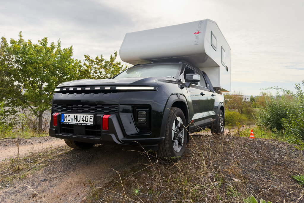 A black SUV with a camper attachment parked on a gravel surface surrounded by greenery.