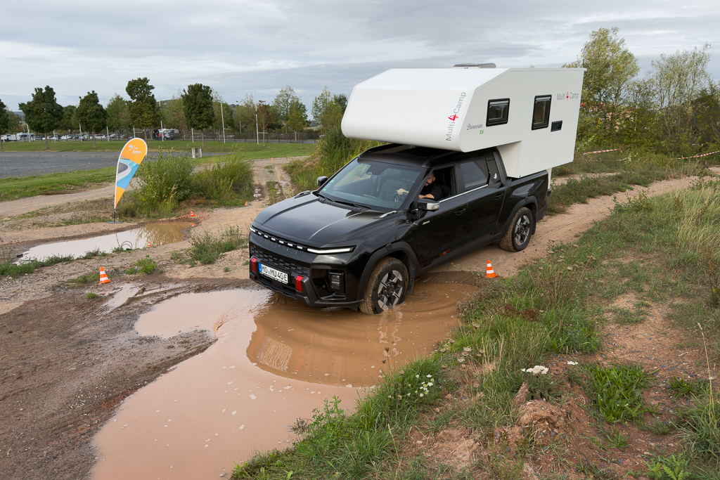A black pickup truck with a white camper on its bed driving through a muddy puddle on an off-road path.