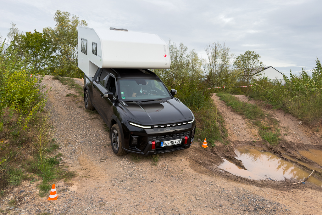 Black pickup truck with a white camper on a gravel off-road trail, surrounded by greenery and a muddy area.