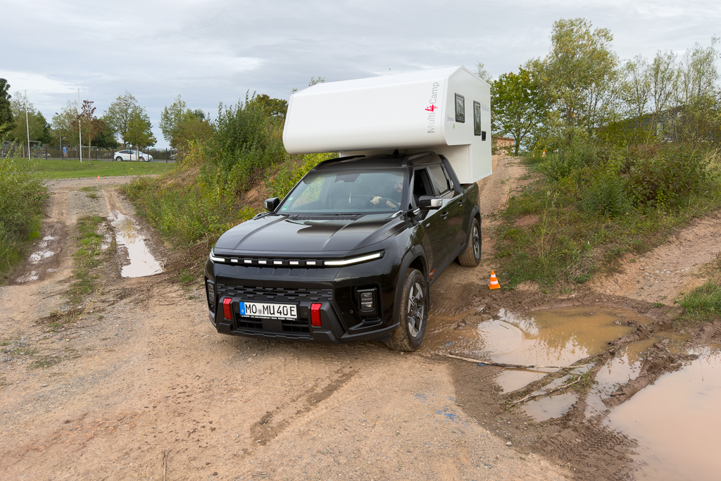 A black pickup truck with a white camper on top parked on a dirt road surrounded by greenery, with puddles visible on the ground.