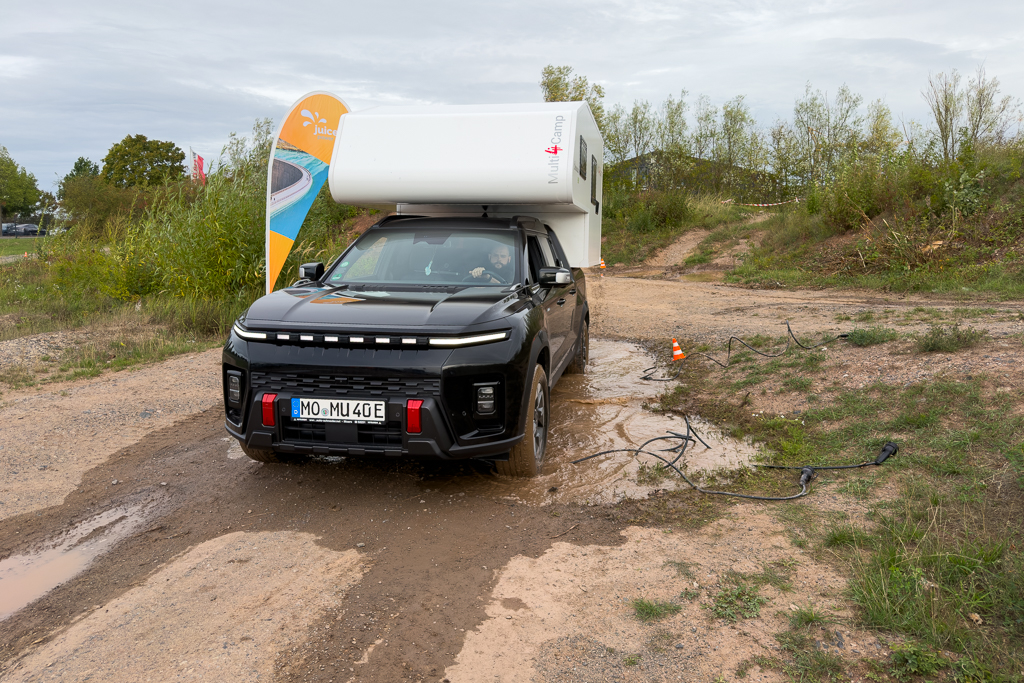 An electric SUV with a camper trailer parked on a muddy outdoor path, surrounded by greenery and a colorful promotional banner.