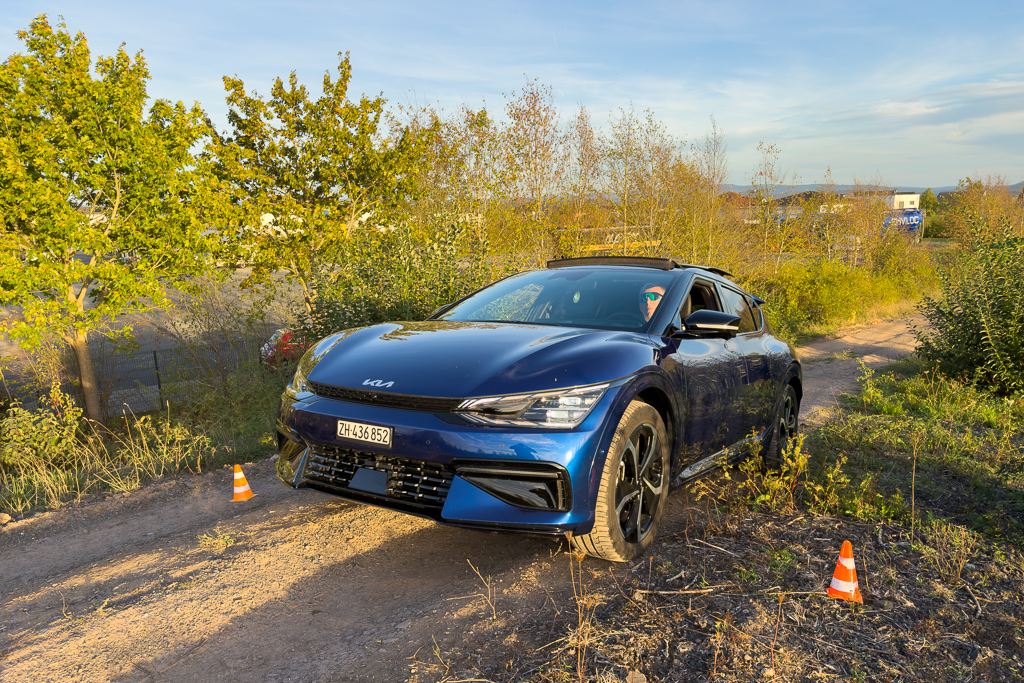 A blue Kia electric vehicle parked on an unpaved road surrounded by trees and shrubs, with traffic cones placed nearby.