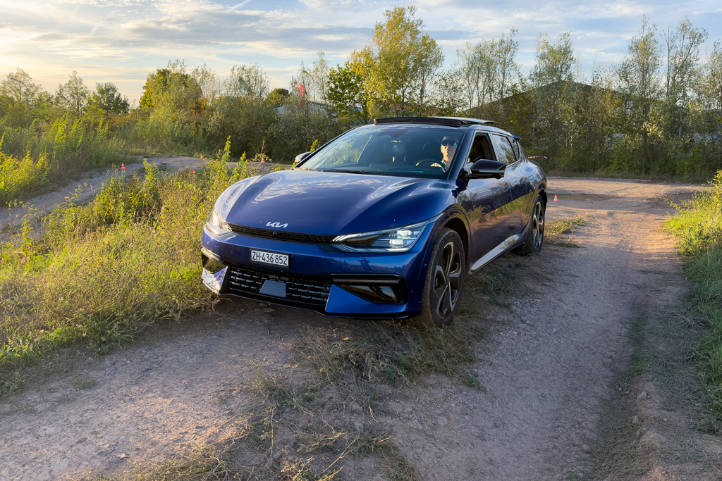 A blue Kia electric vehicle parked on a dirt path surrounded by greenery and trees, with a driver visible inside.