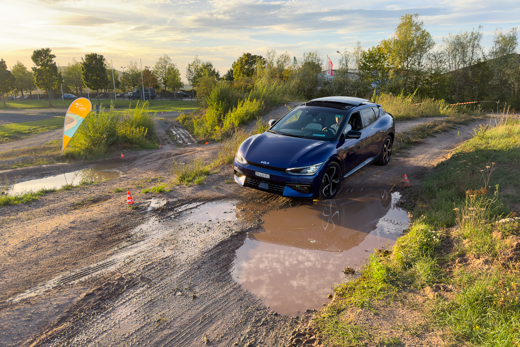 A blue car navigating a muddy off-road course with puddles, surrounded by greenery and trees during sunset.