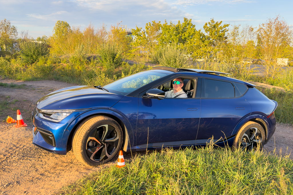 A blue electric car parked on a dirt surface, with a driver wearing sunglasses visible inside. Traffic cones are placed nearby, with greenery in the background under a clear sky.