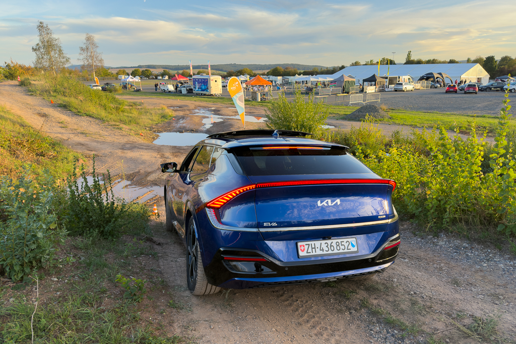 A blue electric car parked on a dirt path near an outdoor event area with tents and vehicles in the background.