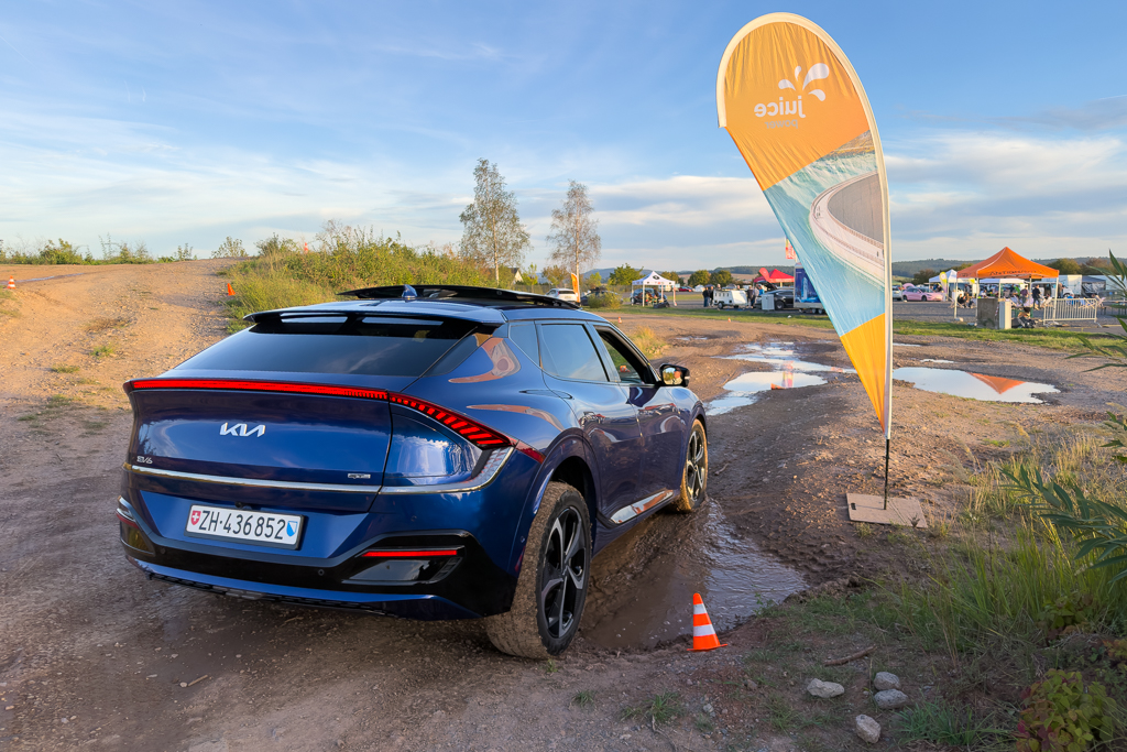 A blue Kia EV6 parked on a dirt road with puddles, near a promotional flag and event setup in the background.