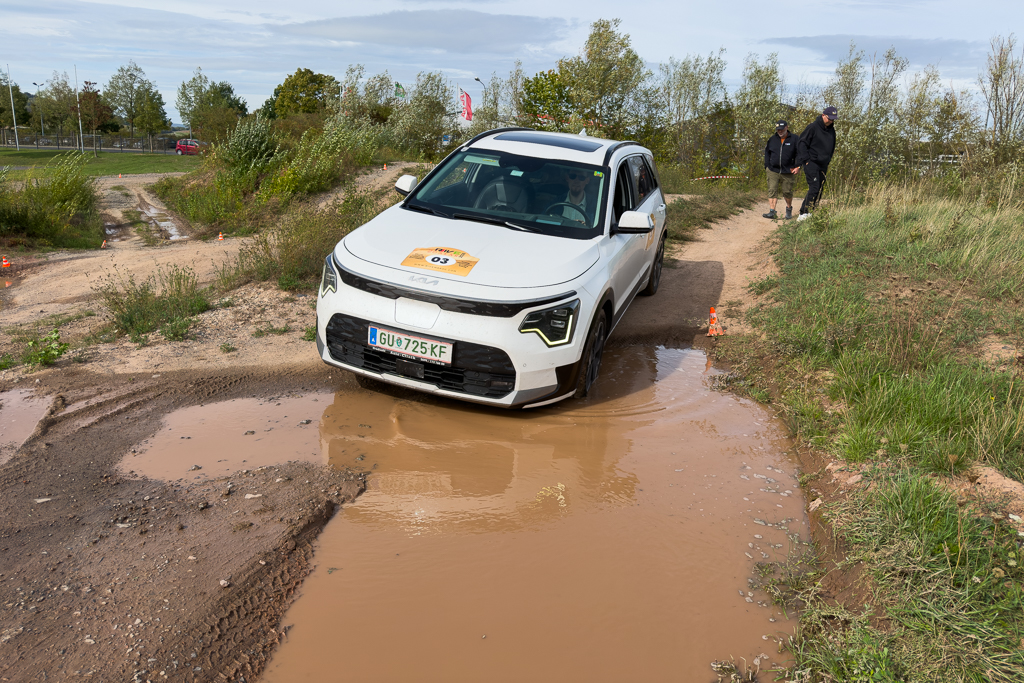 A white SUV navigating through a muddy puddle on an off-road path, with two people walking nearby.