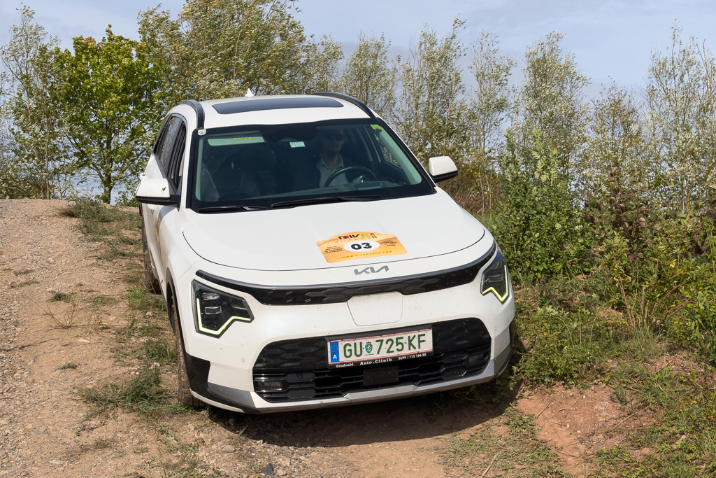 A white Kia SUV parked on a gravel trail with greenery in the background and a competition number plate on the windshield.