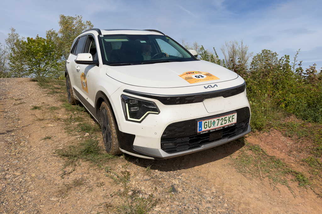 White SUV parked on a gravel path, partially obscured by grass and bushes, with a competition sticker on the windshield.