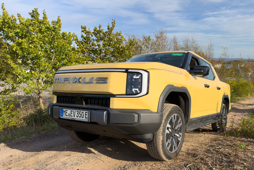 A yellow Maxus electric vehicle parked on a dirt path surrounded by greenery under a blue sky.