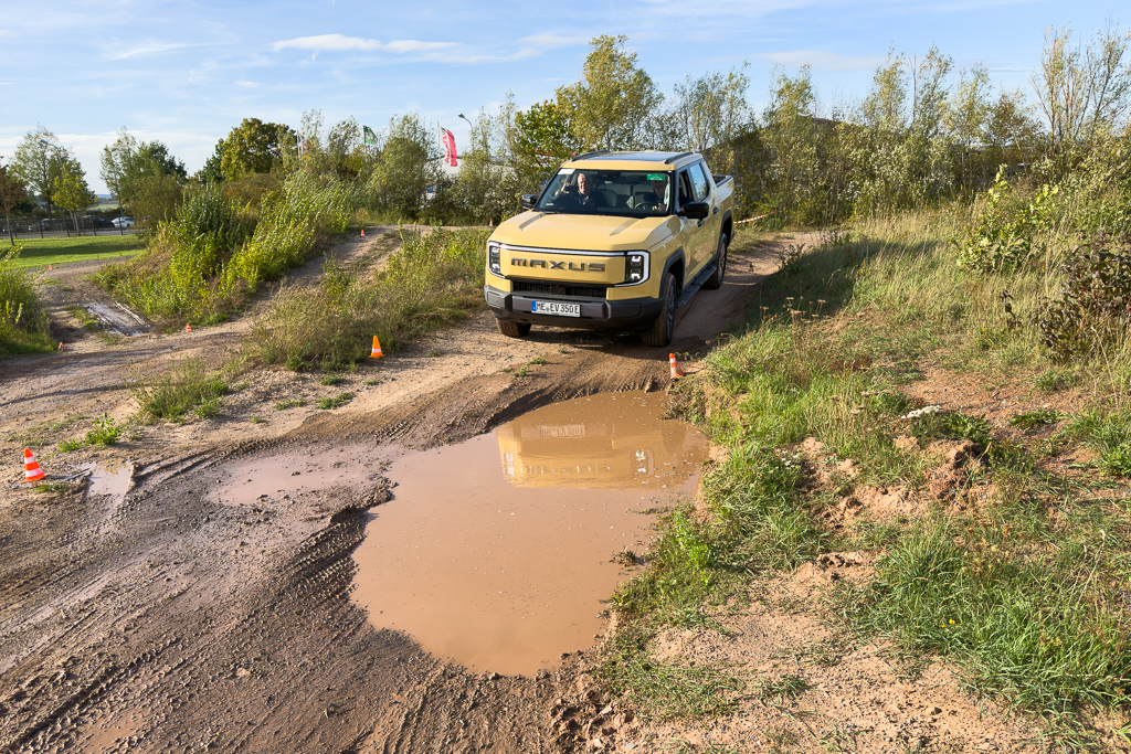A yellow off-road vehicle navigating a muddy terrain with a large puddle, surrounded by greenery and cones marking the path.