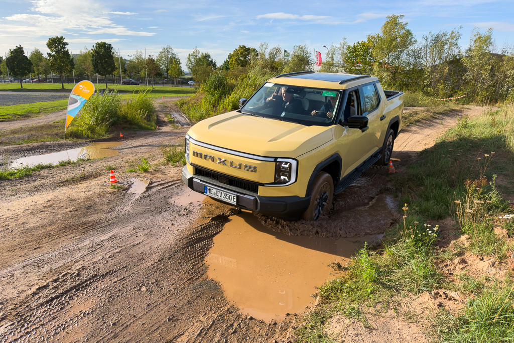 A yellow Maxus pickup truck navigating through a muddy water puddle on a dirt track, surrounded by green grass and trees under a clear sky.