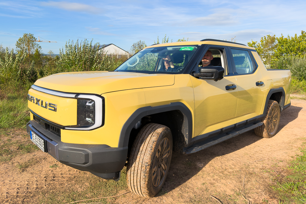 A yellow pickup truck parked on a dirt path surrounded by greenery, with two people visible inside.