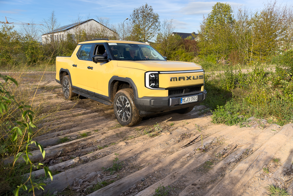A yellow Maxus electric pickup truck parked on a sandy, wooded trail surrounded by greenery.