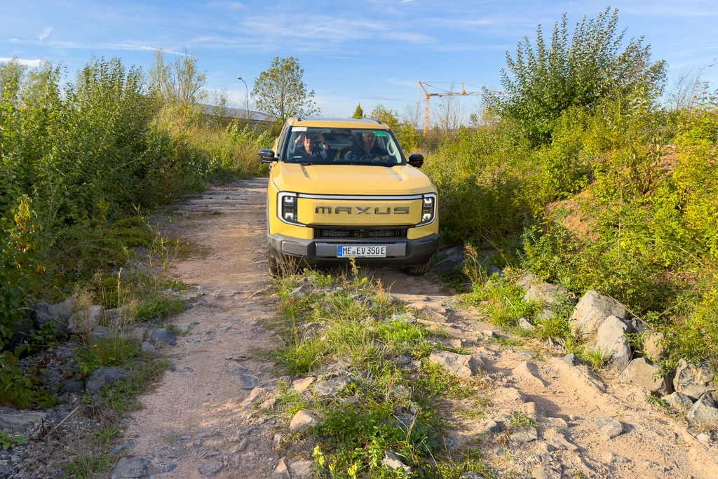 A yellow Maxus vehicle driving on a rugged, overgrown path surrounded by greenery and rocks.