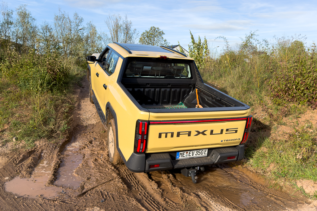 A yellow Maxus T90 electric pickup truck parked on a muddy roadway surrounded by greenery.