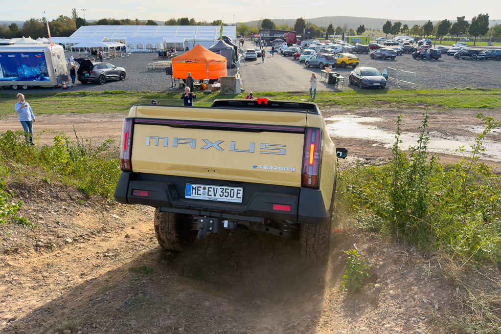 A rear view of a yellow MAXUS Teren 9 electric vehicle climbing a dirt incline, with a parking area and event tents visible in the background.