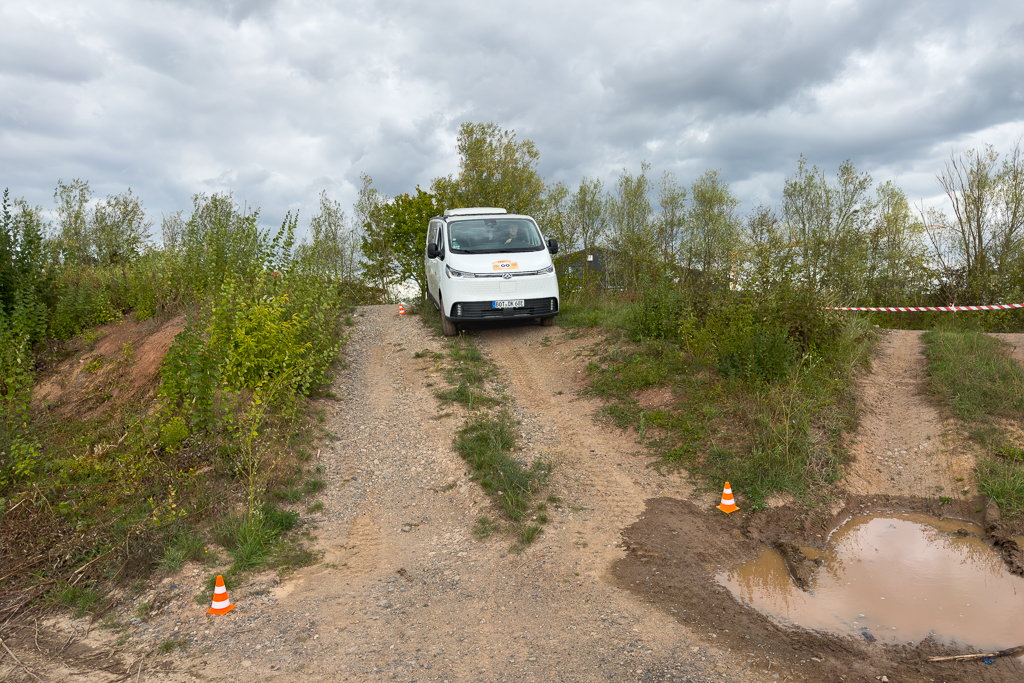 A white van navigating a rough terrain with overgrown grass and a muddy puddle on the right side, under a cloudy sky.