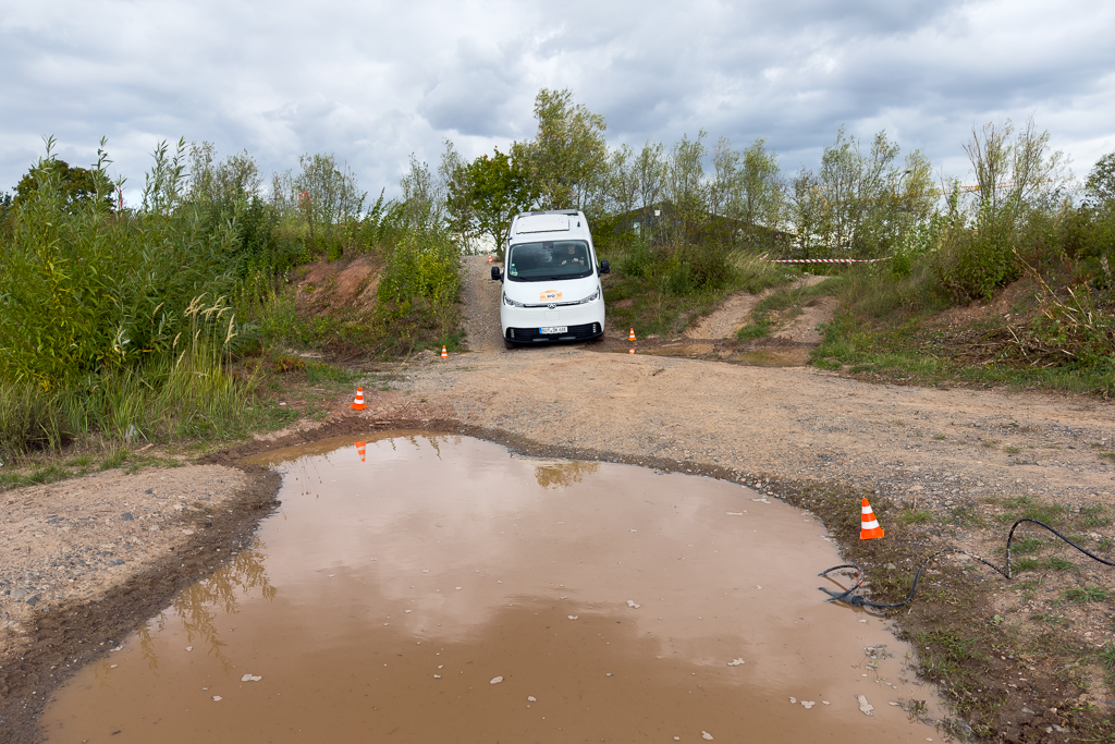 A white delivery van is parked near a large puddle on a gravel road surrounded by greenery and scattered orange traffic cones under cloudy skies.