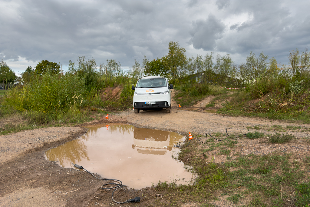 A white van parked near a muddy puddle on a dirt path, surrounded by grass and trees under a cloudy sky.
