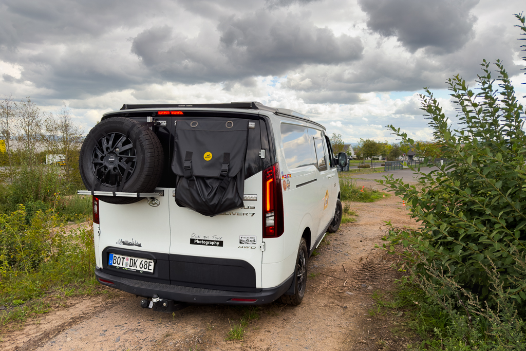 A white camper van parked on a dirt path, featuring a spare tire mounted on the rear door and a black storage bag attached. The background includes green bushes and a cloudy sky.