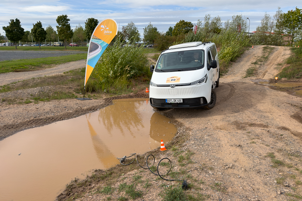 A white electric van driving near a muddy area with standing water, accompanied by a branded flag and orange cones marking the path.