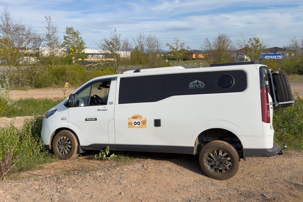 A white van with off-road capabilities parked on a gravel surface, partially surrounded by greenery. The vehicle features a custom design with a logo and is equipped with specialized tires.