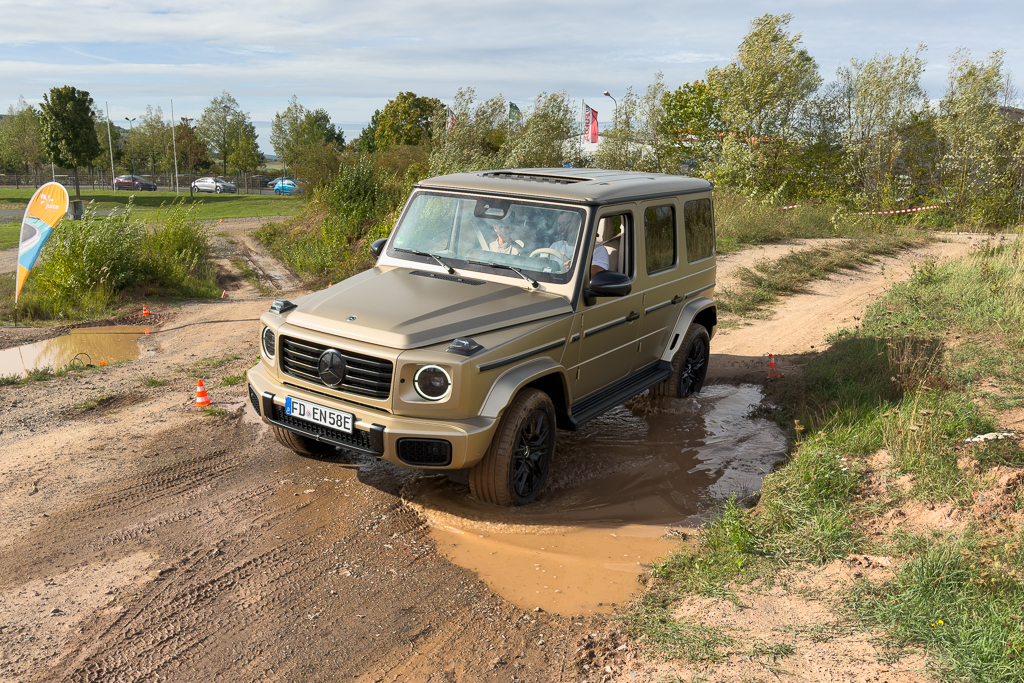 A beige Mercedes-Benz G-Class off-roading vehicle navigating through a muddy puddle on a dirt path surrounded by greenery.