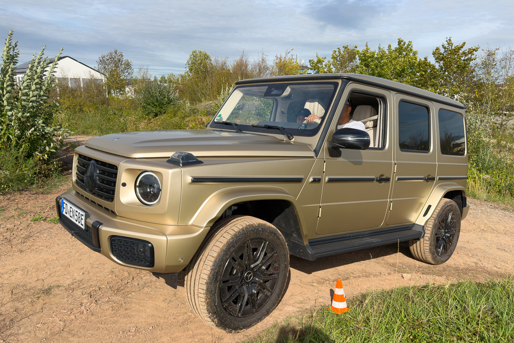 A luxury SUV, specifically a Mercedes-Benz G-Class, parked on a dirt path surrounded by greenery. The vehicle is beige with black rims, and a driver is inside, navigating through an off-road environment.