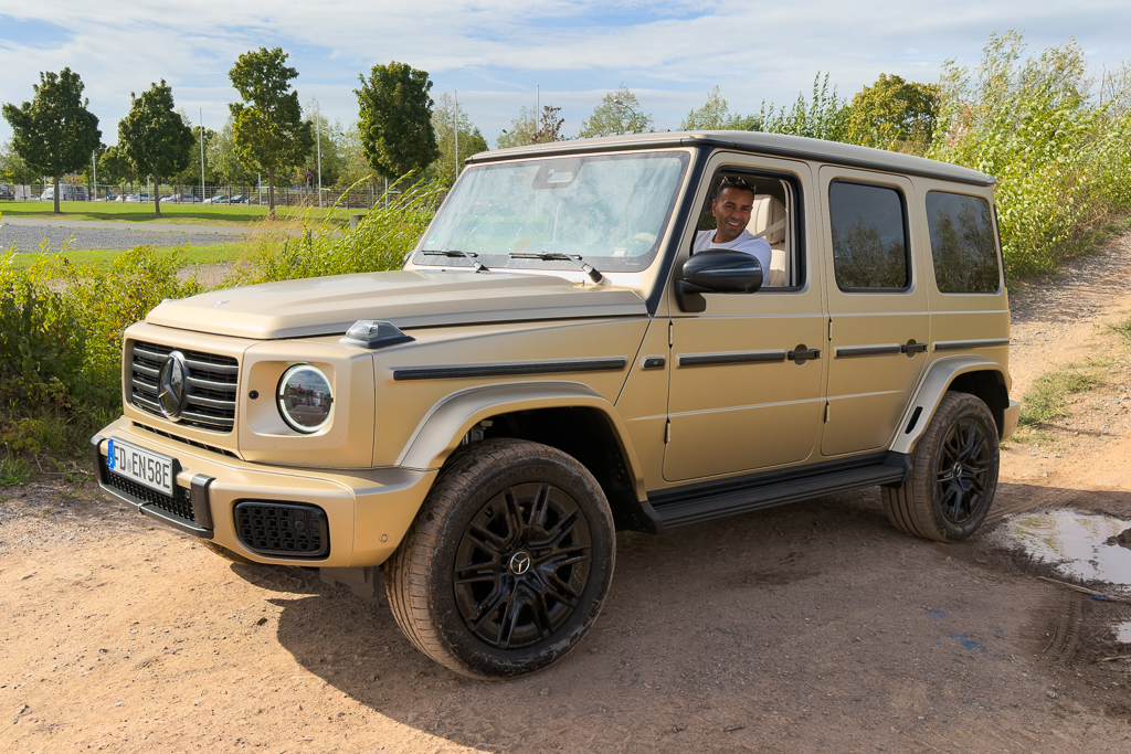 A beige Mercedes G-Class SUV parked on a dirt road with a smiling driver inside. The background features greenery and trees.