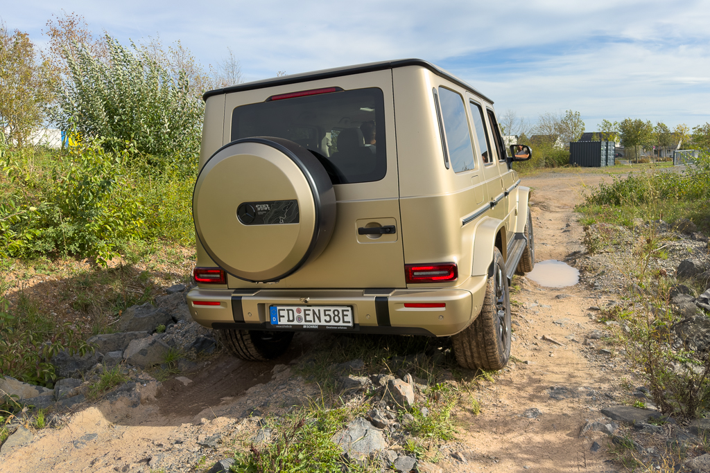 A rear view of a beige off-road vehicle on a rocky terrain, surrounded by vegetation and a clear sky.