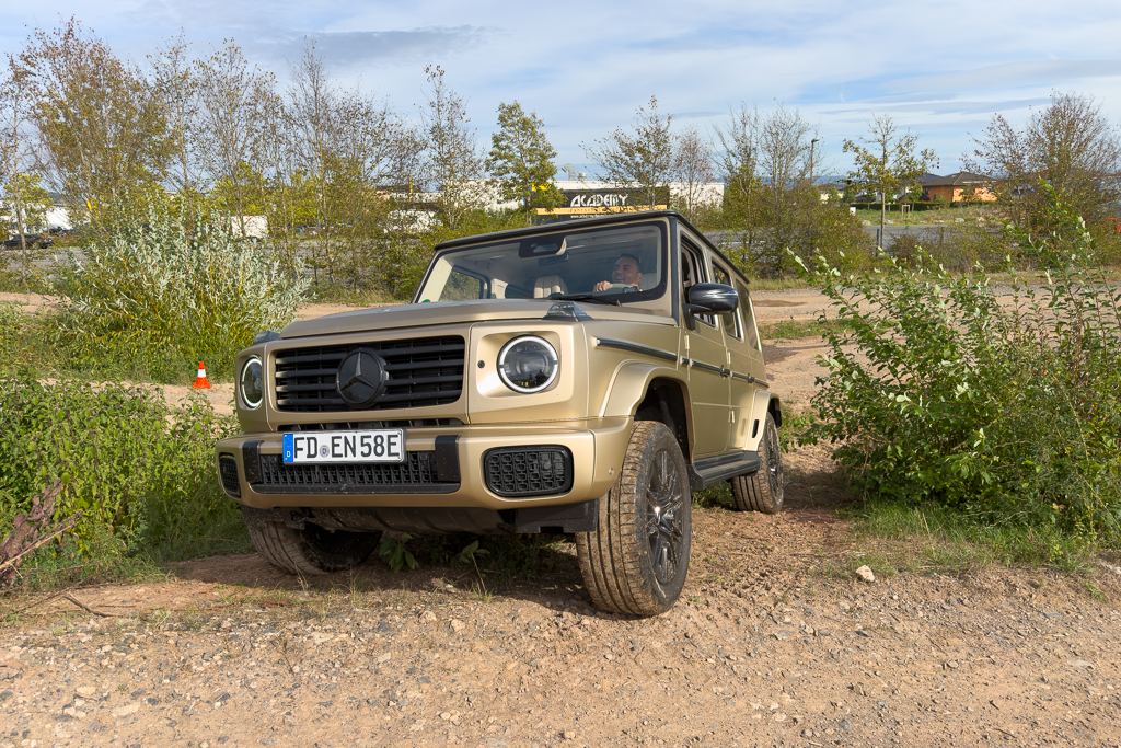 A beige Mercedes-Benz G-Class SUV parked on rugged terrain surrounded by greenery and trees.