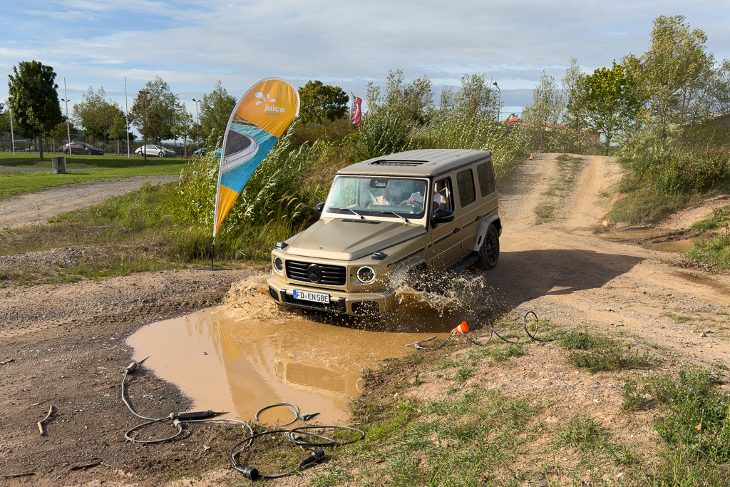 A beige off-road vehicle driving through a muddy puddle with water splashing, surrounded by greenery and a banner in the background.