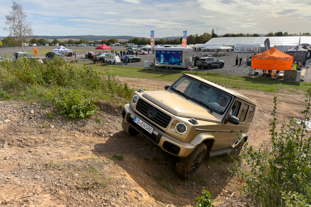 A beige off-road vehicle navigating a steep dirt incline at an outdoor event, with tents and other cars visible in the background.