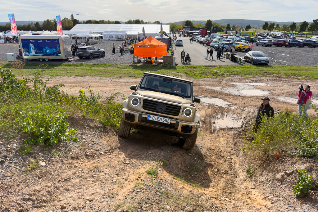 A beige off-road vehicle navigating a rugged dirt incline, surrounded by a grassy area and event tents in the background, with spectators observing and taking photographs.