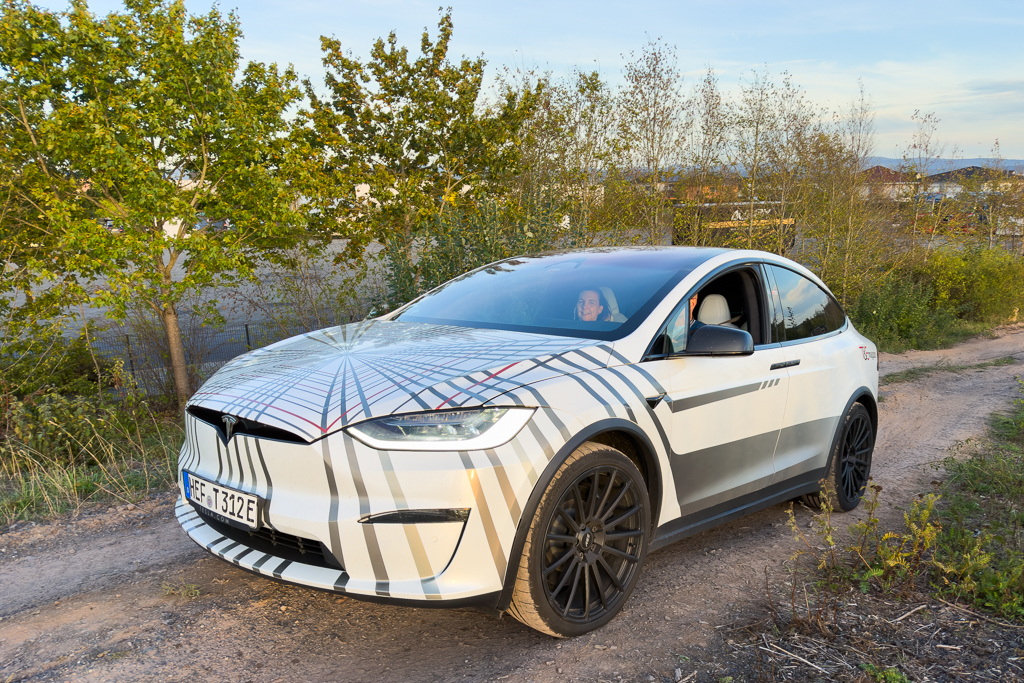 A Tesla Model X parked on a dirt path surrounded by greenery, featuring a unique geometric design on its exterior, with a driver visible inside.