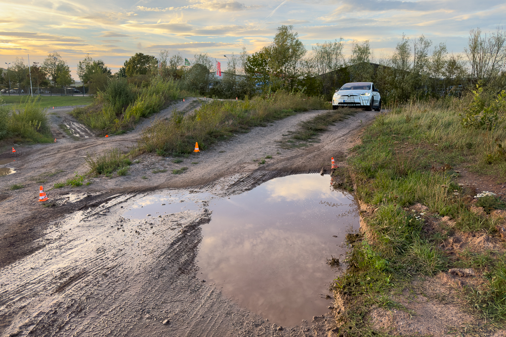 An off-road vehicle navigating a dirt track with puddles, surrounded by greenery and a scenic sky during sunset.