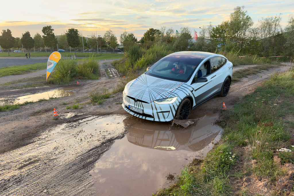 A white electric SUV navigating through a muddy path with puddles, surrounded by greenery and a sunset sky.