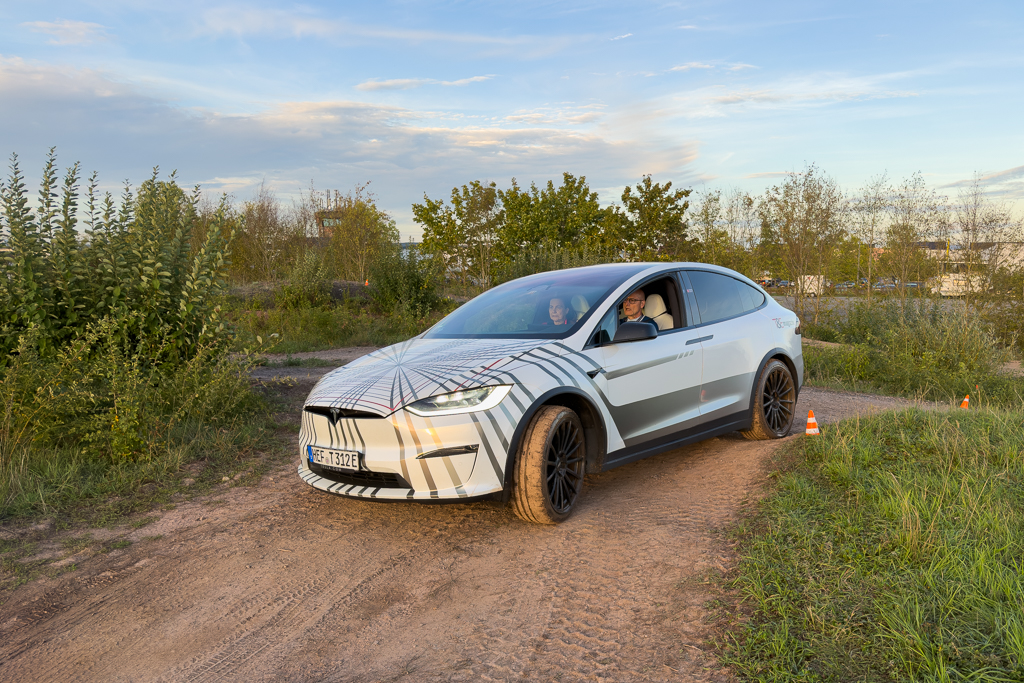 A camouflaged electric SUV driving on a dirt track in a natural setting, with two occupants visible inside the vehicle.
