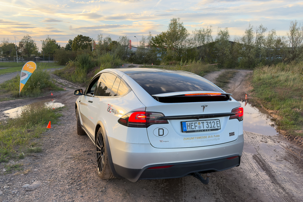 A white Tesla Model X parked on a dirt road with puddles, near a flag indicating an event, under a sunset sky.