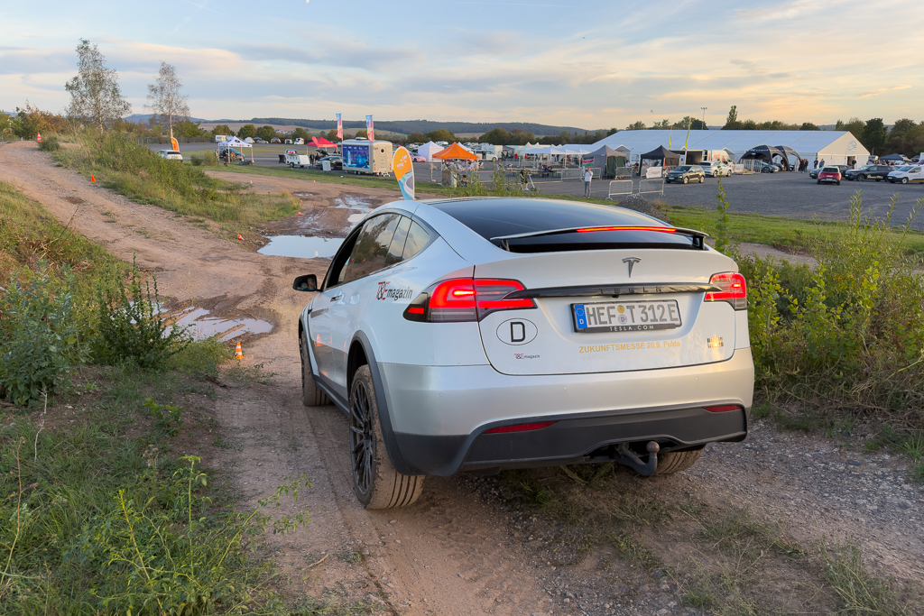 A Tesla Model X parked on a dirt road near an event location, with tents and banners visible in the background.