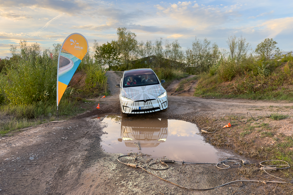 A vehicle with a distinctive pattern navigating a dirt track with a puddle, accompanied by a decorative flag labeled 'Juice', surrounded by greenery and a cloudy sky.