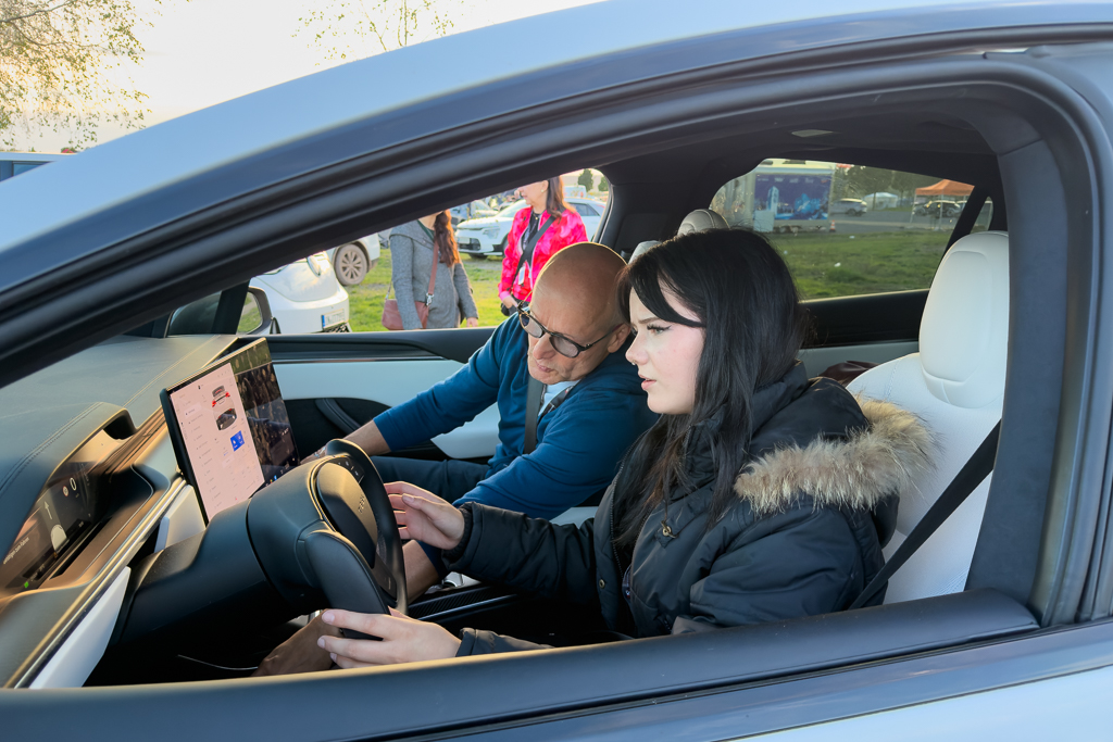 A man and a woman inside a car, examining the touchscreen display. The woman is focused on the screen while the man gestures, providing guidance. The setting appears to be an outdoor event with cars in the background.