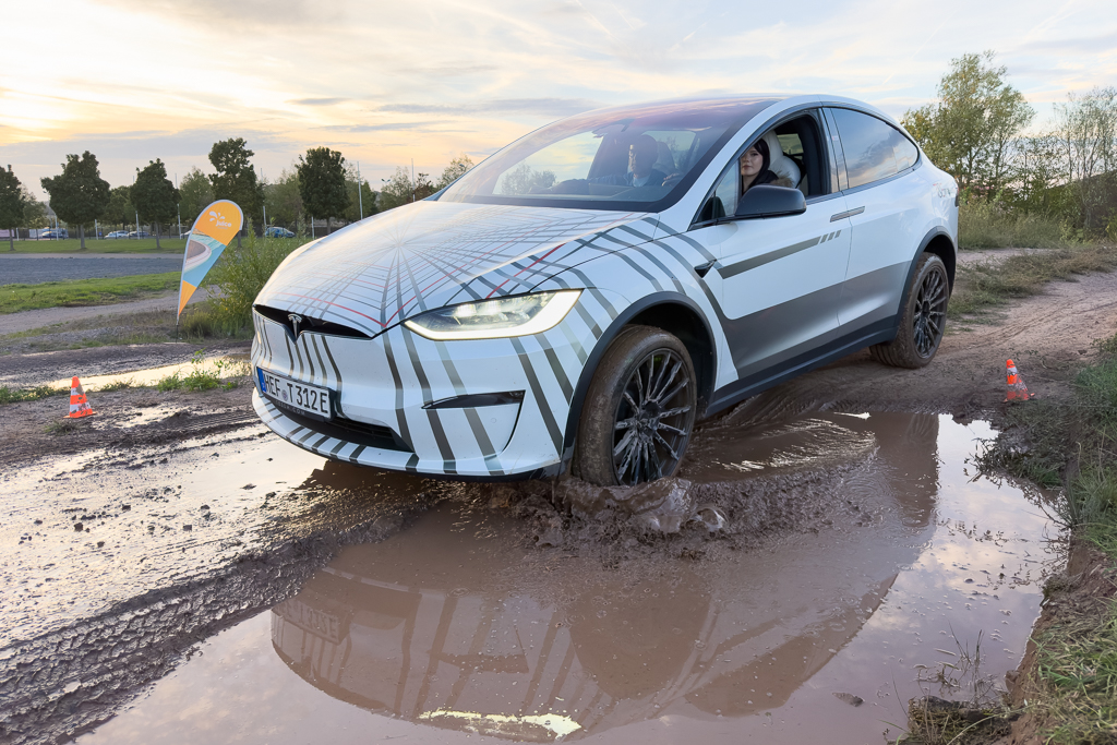 A Tesla Model X driving through a muddy puddle during an outdoor demonstration, with trees and a flag in the background.